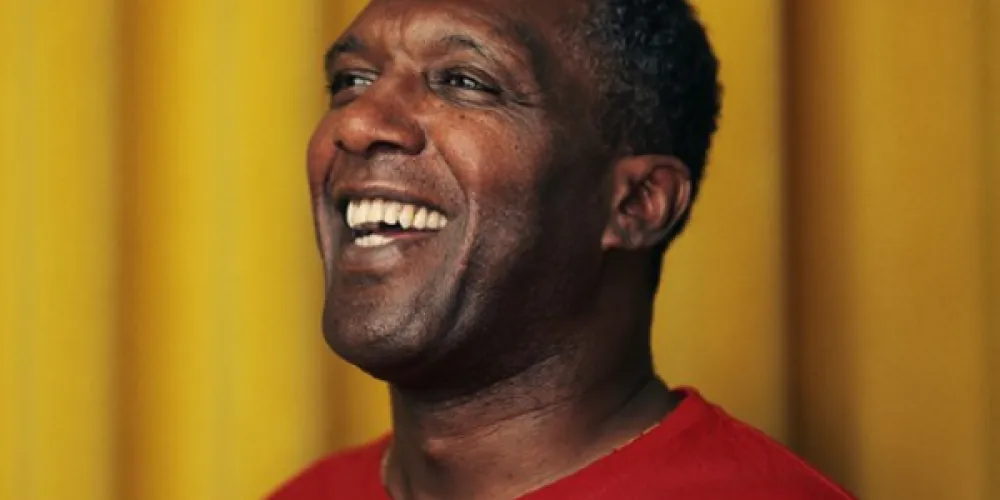 Photo of Lemn Sissay, smiling in a red shirt against a yellow backdrop.